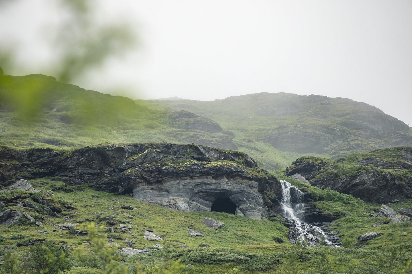 Norwegian river winding past cave entrance in lush green mountains, overcast sky.