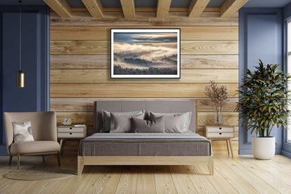 Framed print of Forest valley shrouded in mist, sunrise casts delicate yellow hues, on a wooden wall in a bedroom.