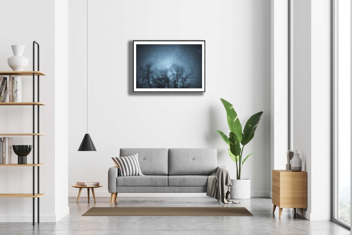 Long-exposure winter storm photo, swaying trees against starry sky, hung on white wall above sofa.