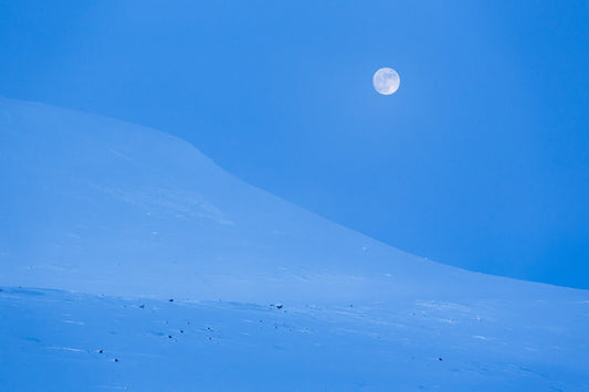 Tranquil Arctic landscape photo captured during blue hour, with full moon illuminating snow-capped mountains.