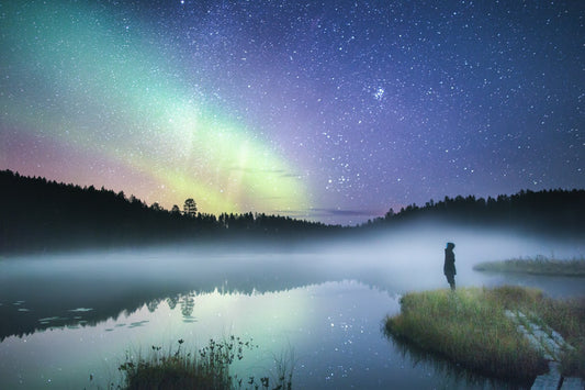 A person standing on the shore of a foggy forest pond on an autumn night, reflecting the Northern Lights and stars.
