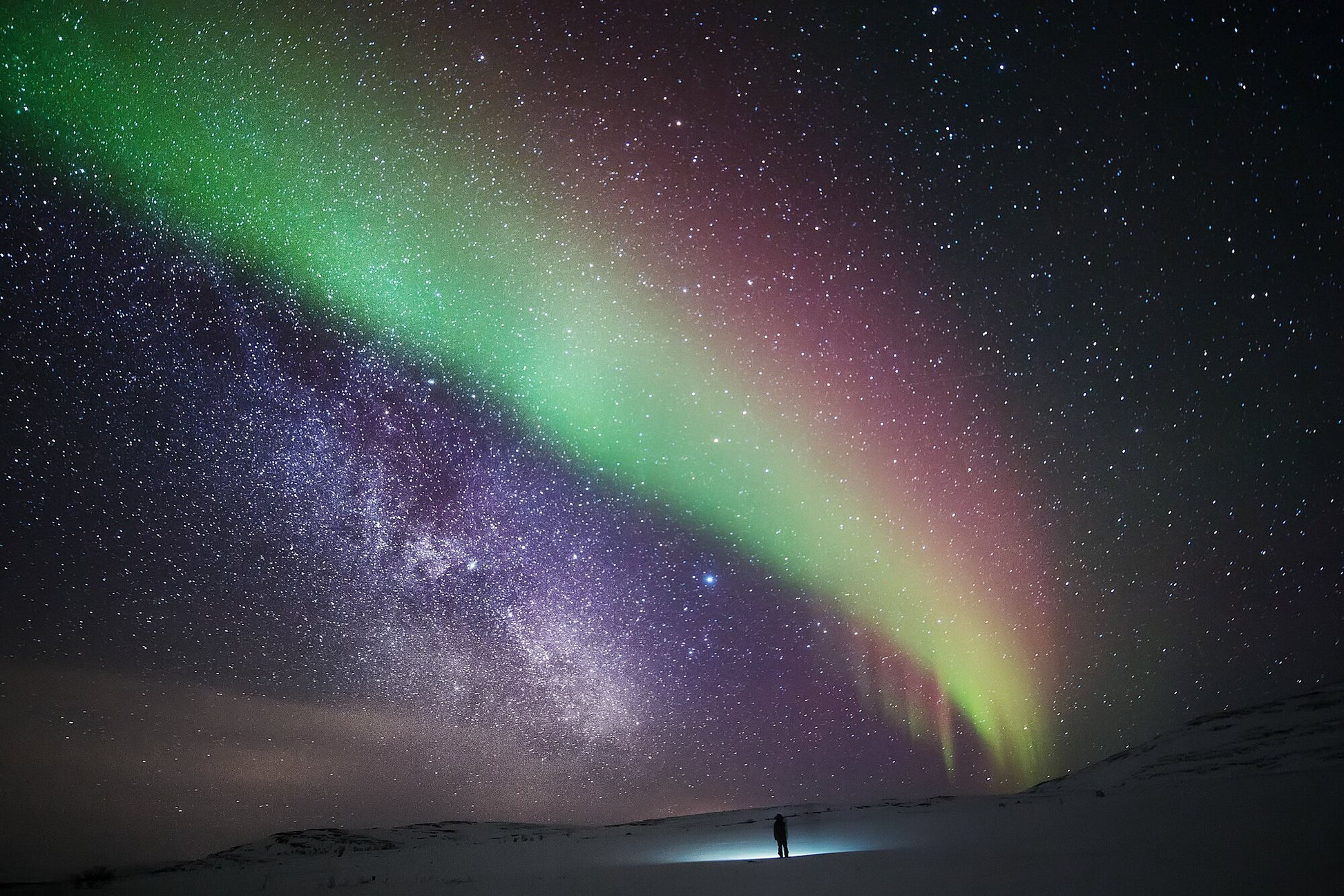 A person stands in the Arctic wilderness, gazing at the aurora borealis and the stars creating an otherworldly spectacle.
