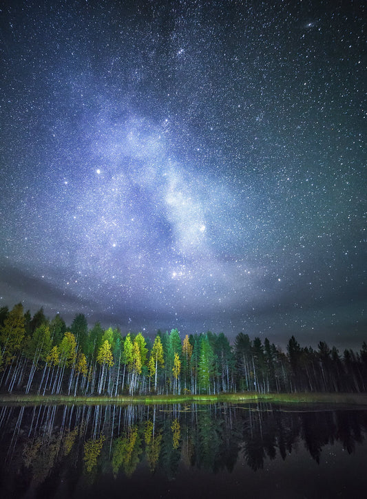 Autumn forest reflected on tranquil water, stars and Milky Way visible in the night sky.