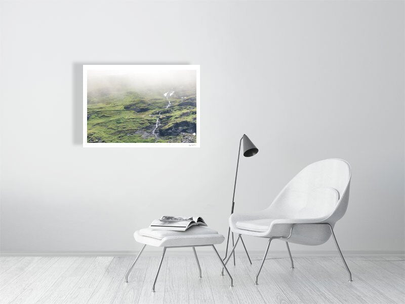 Photo of thin stream descending green Norwegian mountain with cloud-covered peak and unmelted snow, white living room wall.