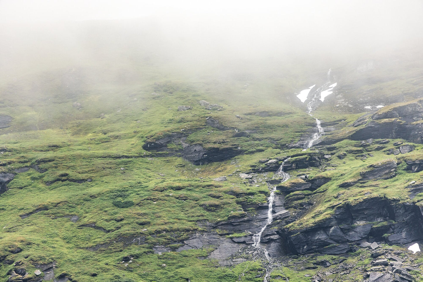 Thin stream flows down green Norwegian mountain with cloud-covered peak and unmelted snow.