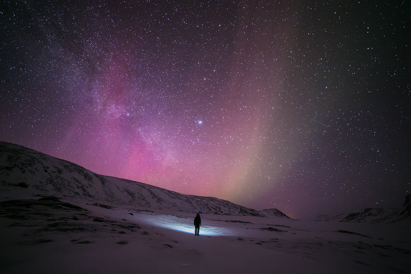 Person standing alone in Arctic wilderness during midwinter, gazing at stars and pink aurora borealis.