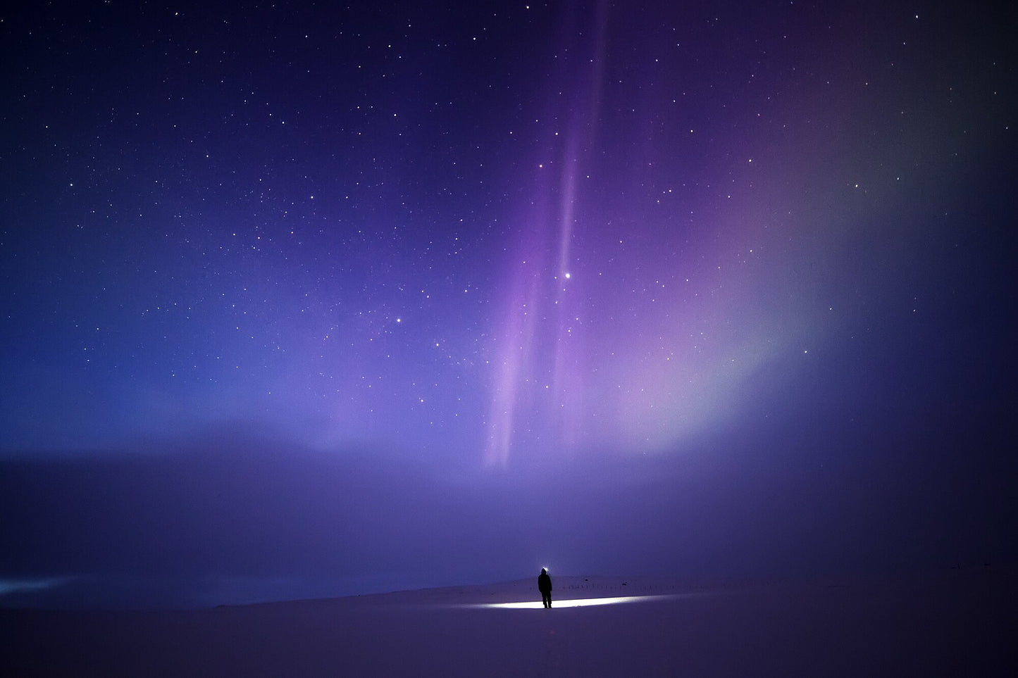 Person stands alone in Arctic wilderness under aurora borealis and fainting clouds.