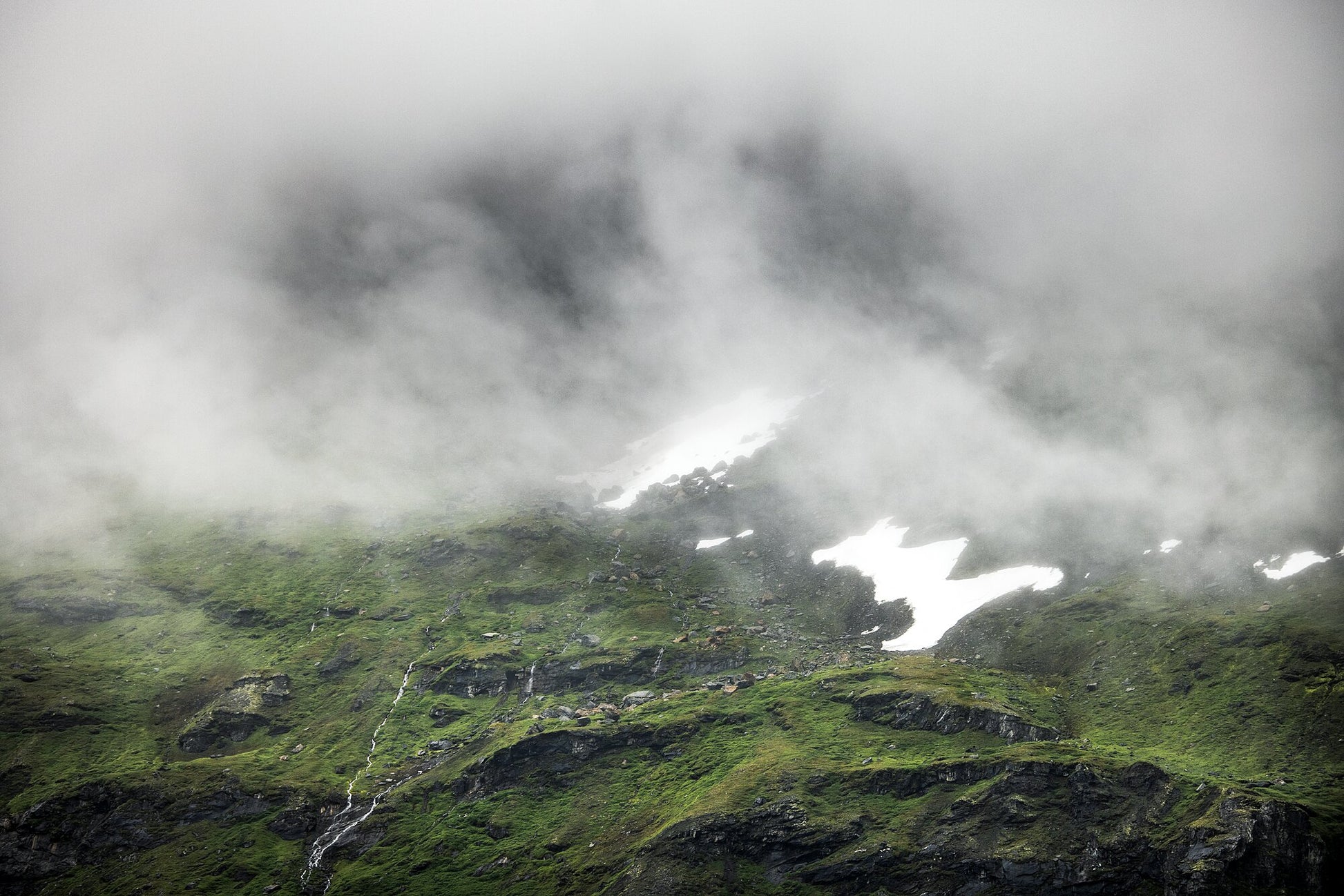 Cloud-shrouded Norwegian mountain peak with meltwater streams and snowy patches.