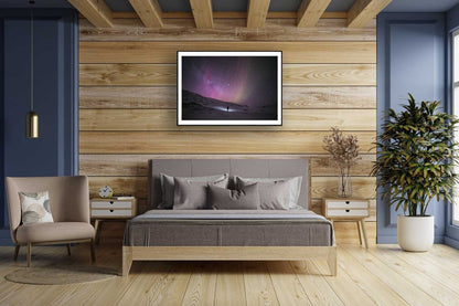 Framed photo of person gazing at stars in Arctic wilderness, pink aurora borealis, wooden bedroom wall.