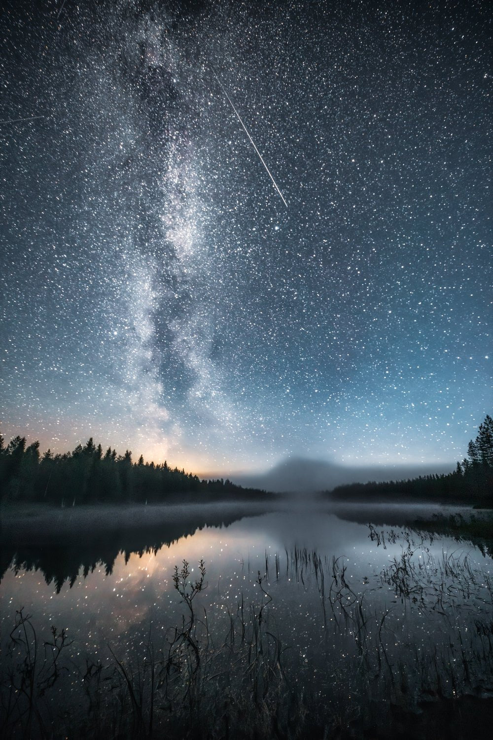 Night sky photo of Milky Way and stars mirrored in lake with shooting star, capturing the feeling of a northern autumn night.