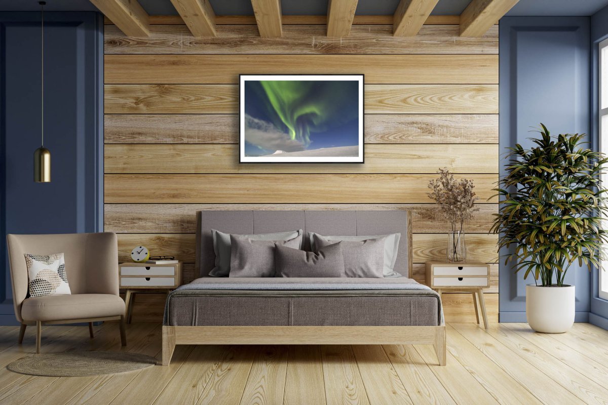 Art print of mountains, moonlight & Northern Lights, black-framed on wooden wall above bed.
