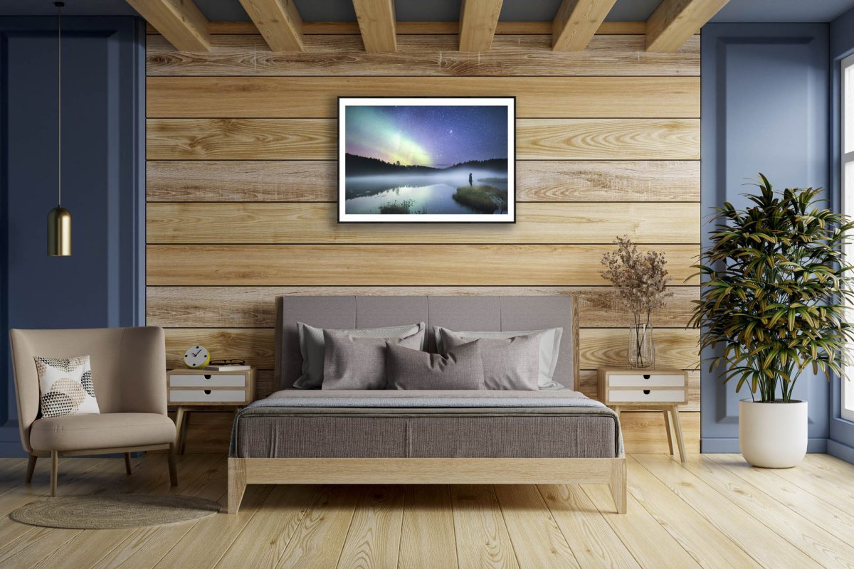 Fine art print of a person in the forest at night, reflecting aurors and stars, black-framed on a wooden wall in the bedroom.