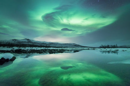 Arctic landscape with Northern Lights reflecting in lake and snowy mountains in the distance.