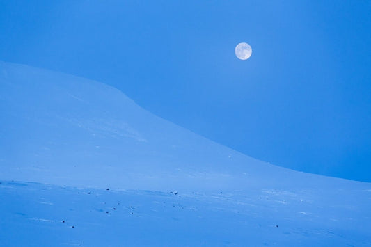 Tranquil Arctic landscape photo captured during blue hour, with full moon illuminating snow-capped mountains.