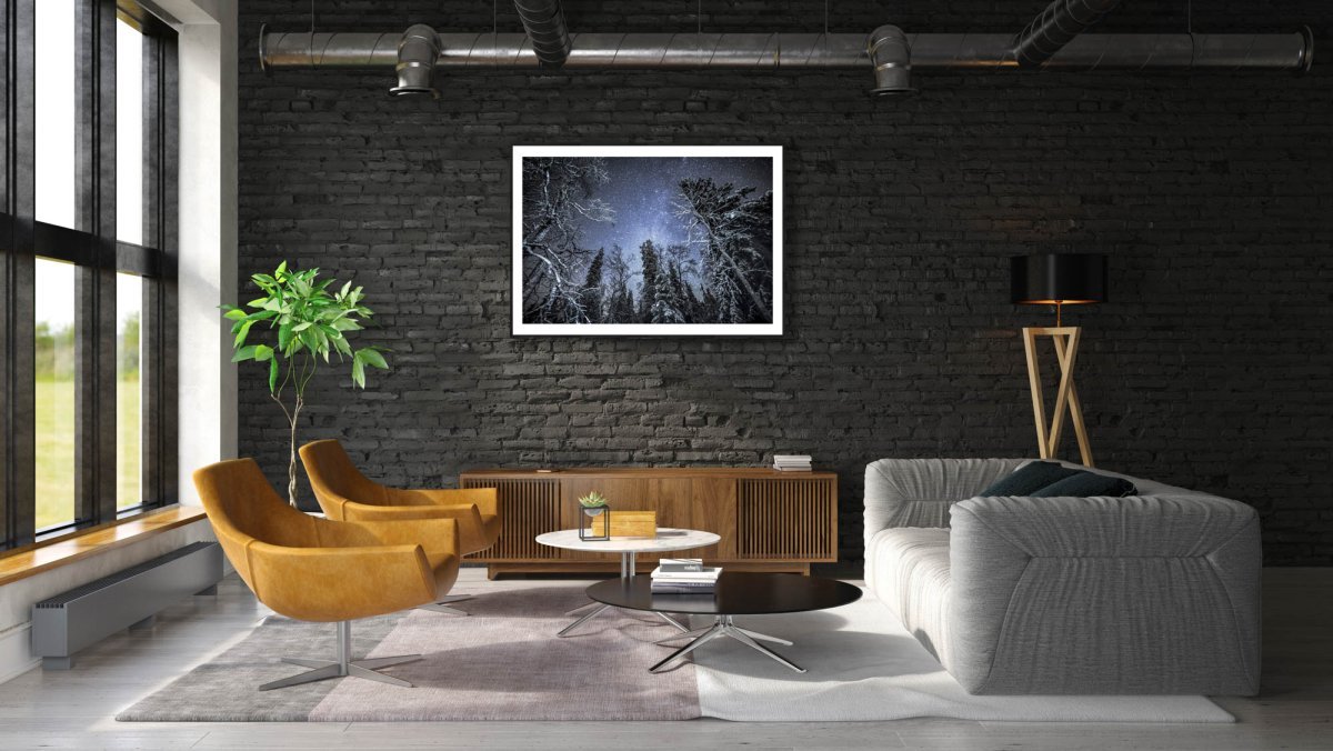Framed illuminated trees in winter forest under starry sky, hung on black brick wall above cabinet.