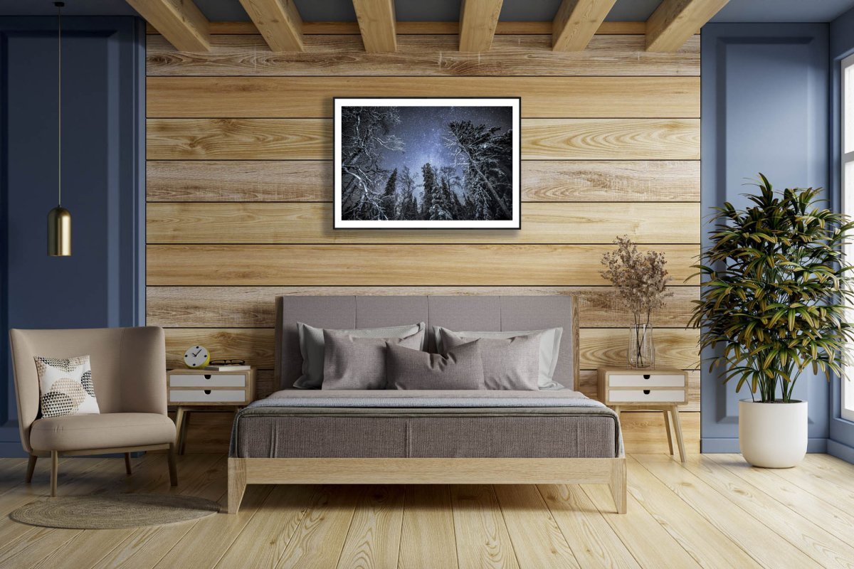 Framed illuminated trees in winter forest under starry sky, hung on wooden wall above bed.