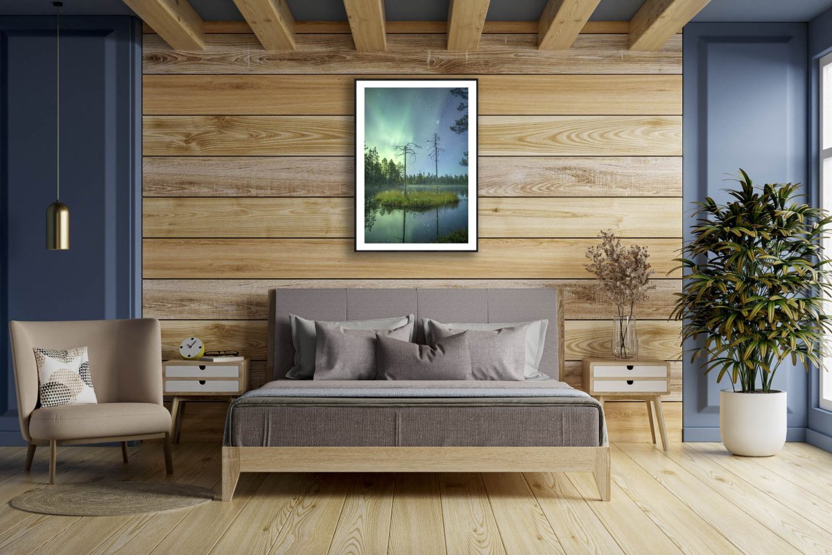 Framed photo, forest pond with island and pines reflecting Northern Lights and starlit sky, wooden bedroom wall above bed.