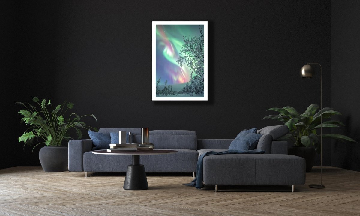 Fine art photography print of Aurora borealis over snowy forest, black-framed, on black wall in modern living room above sofa.