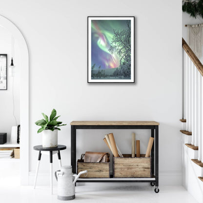 Fine art photography print of Aurora borealis over snowy forest, black-framed, on white living room wall above desk.
