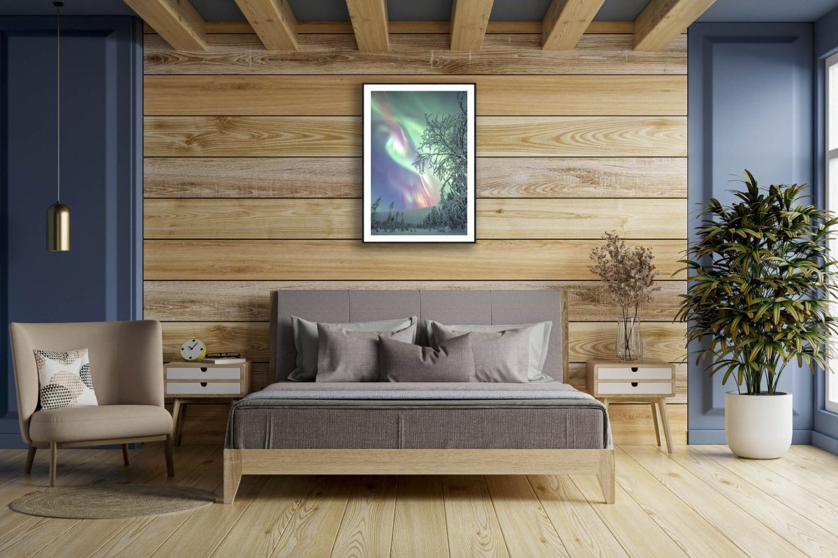 Fine art photography print of Aurora borealis over snowy forest, black-framed, on wooden bedroom wall above bed.