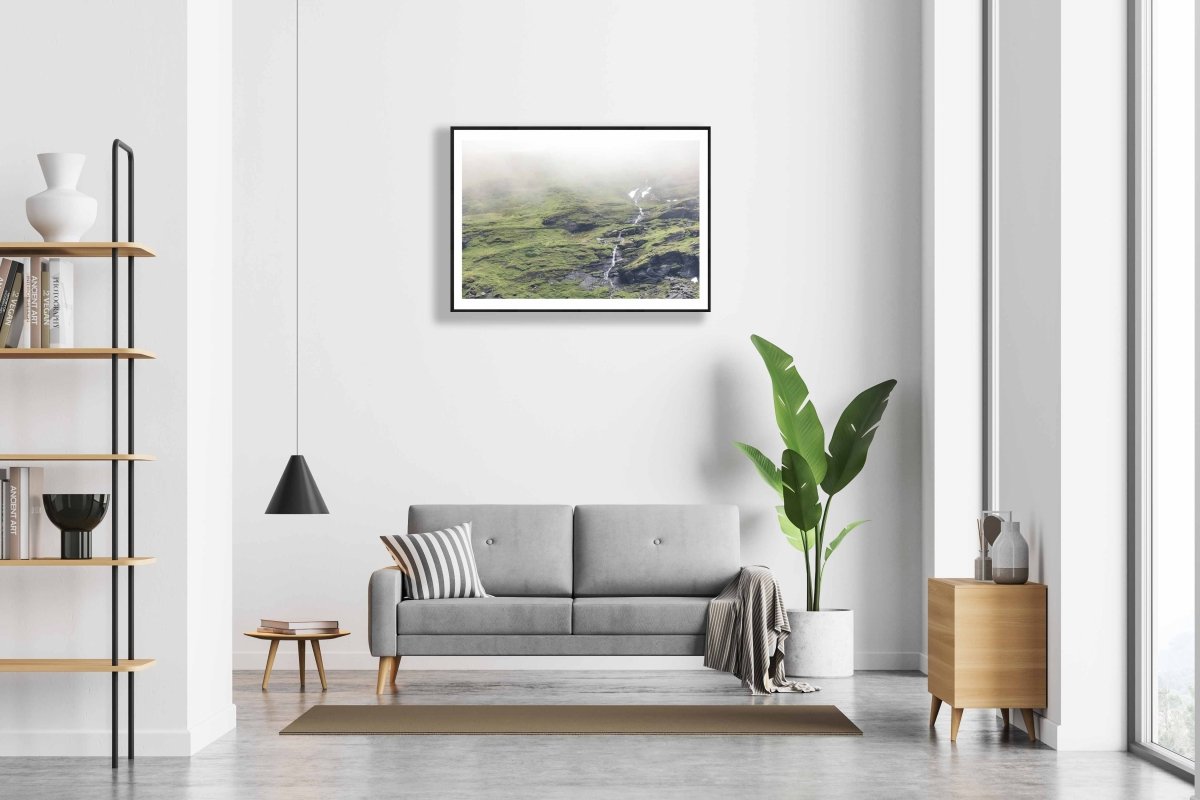 Framed photo of thin stream descending green Norwegian mountain with cloud-covered peak and snow, white living room wall.