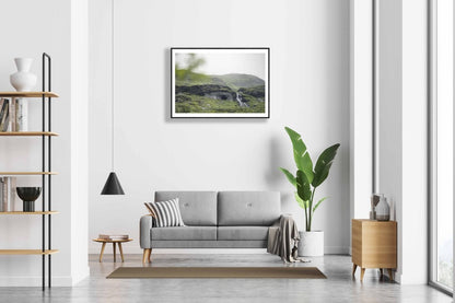 Framed photo of Norwegian river winding past cave entrance in lush green mountains, white living room wall.